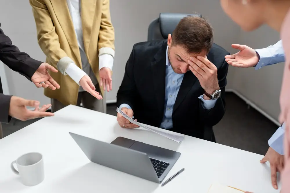 A imagem contém um trabalhador ao meio, com as mãos na cabeça, com expressão de constrangido, ao lado de 3 pessoas com mãos direcionadas a ele como se estivessem fazendo alguma cobrança.