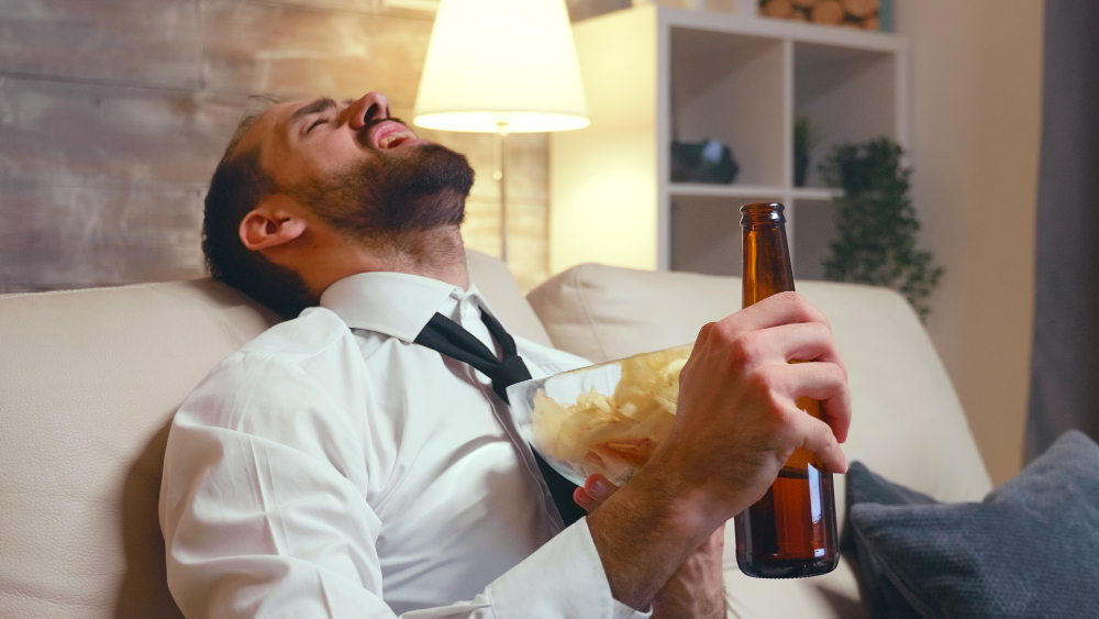 A imagem contém um homem, de barba, camisa branca, gravata preta, sentado em um sofá branco, com uma cerveja na mão e aparentemente embriagado.