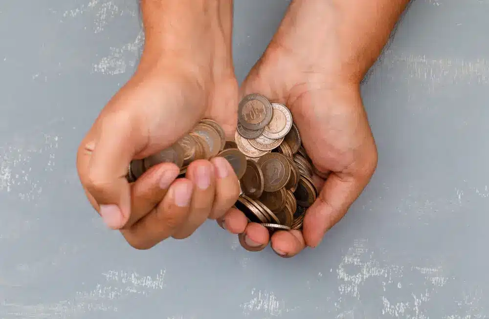 A imagem contém 2 mãos segurando diversas moedas. O fundo é similar a um cão de cimento.